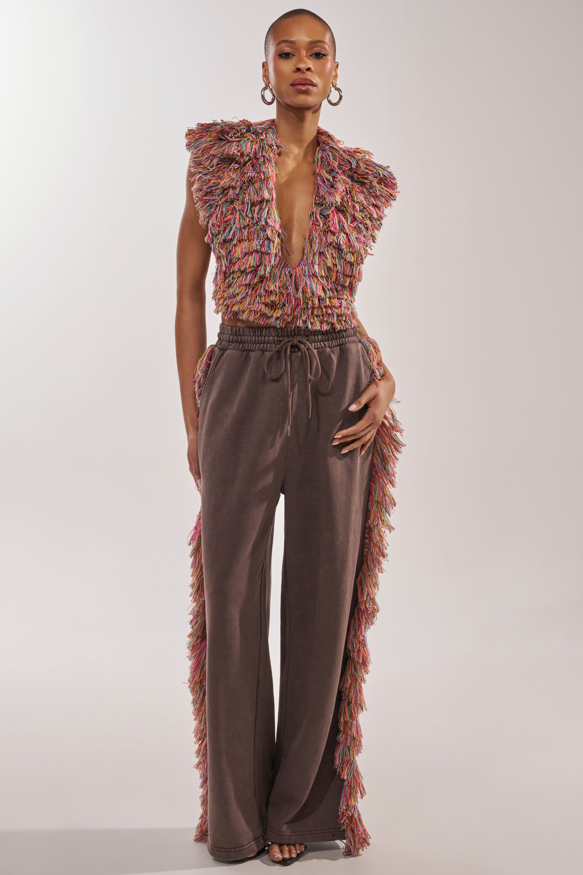 A person stands confidently in THE ONE FRINGE JOGGER—wide-leg brown sweatpants with a drawstring and rainbow fringe along the sides—styled with a sleeveless, multicolored fringed top for perfect festival streetwear. 8