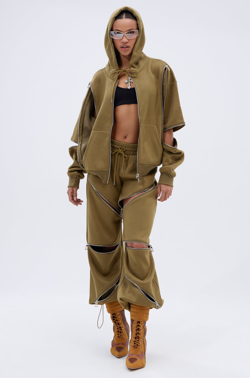 A model wears khaki green CALI MULTI ZIPPER SWEATPANT with a matching hoodie, black crop top, brown lace-up heeled boots, and clear futuristic glasses while standing against a plain white background.
