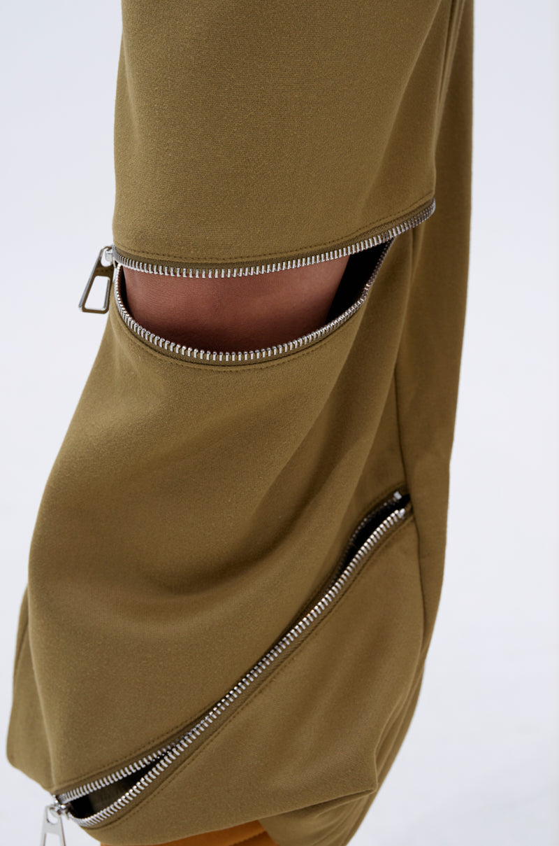 A close-up of the CALI MULTI ZIPPER SWEATPANT in olive green, featuring two horizontal zippers on the leg—one partly unzipped to show skin—crafted from a smooth, slightly draped fabric.