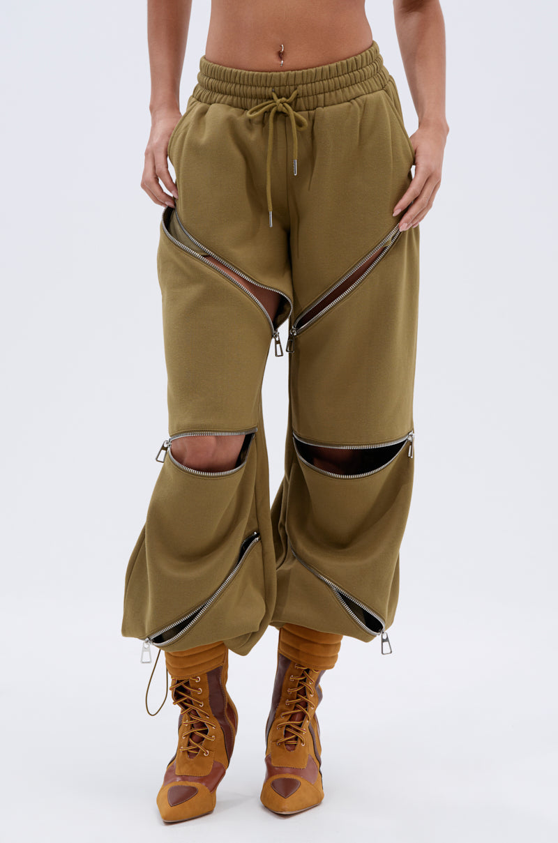 Someone wears the CALI MULTI ZIPPER SWEATPANT, featuring multiple horizontal zippers partially open at the thighs and knees, paired with brown lace-up boots. The background is plain white.