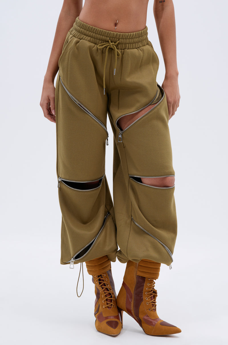 Wearing the CALI MULTI ZIPPER SWEATPANT in olive green with multiple diagonal zippers, paired with orange and brown lace-up boots. Midriff is exposed, showing a belly button piercing.