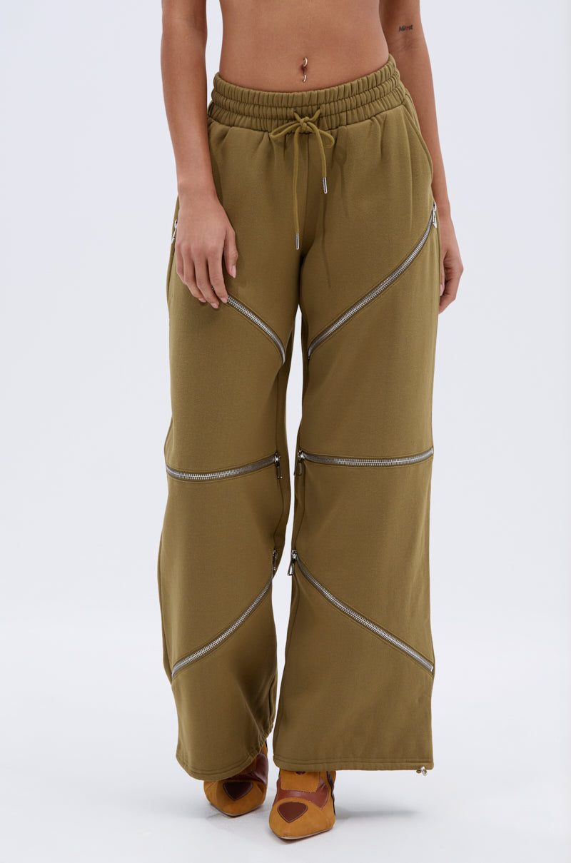 A person wears CALI MULTI ZIPPER SWEATPANT in olive green, featuring diagonal silver zippers and an elastic drawstring waist, paired with orange shoes. The relaxed-fit sweatpants are shown in a photo cropped from chest down.