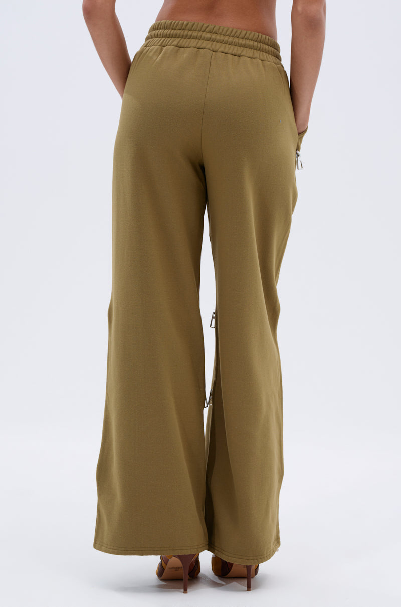 A person is seen from behind wearing CALI MULTI ZIPPER SWEATPANT, featuring a loose, relaxed fit and olive-green color with an elastic waistband, standing against a plain white background.