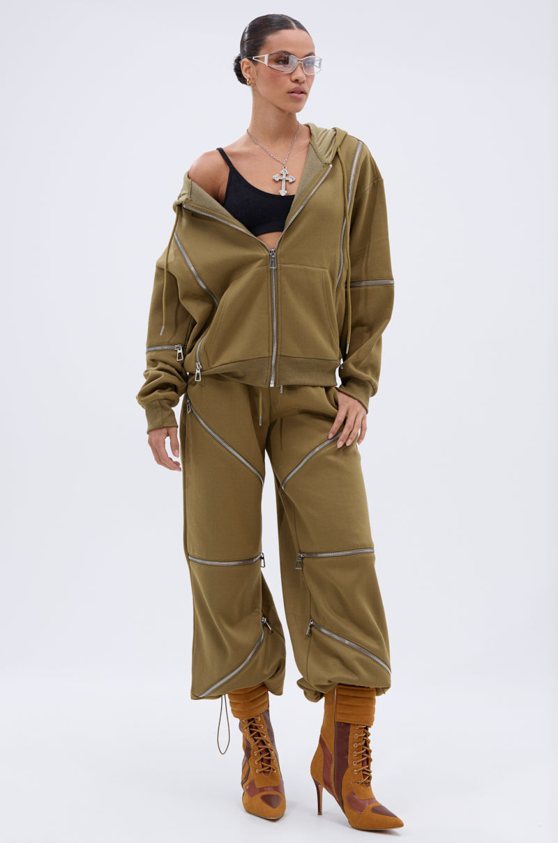 A woman stands against a plain background wearing the CALI MULTI ZIPPER SWEATPANT and an oversized olive green zip-up jacket, paired with a black crop top, tan lace-up heeled boots, clear glasses, and a large cross necklace.