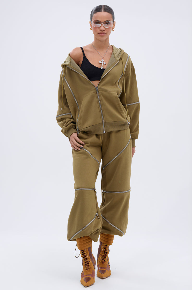 A woman stands confidently against a white background in the CALI MULTI ZIPPER SWEATPANT, an olive green relaxed-fit sweatpants tracksuit with white piping, paired with a black bralette, brown lace-up boots, sunglasses, and cross necklaces.