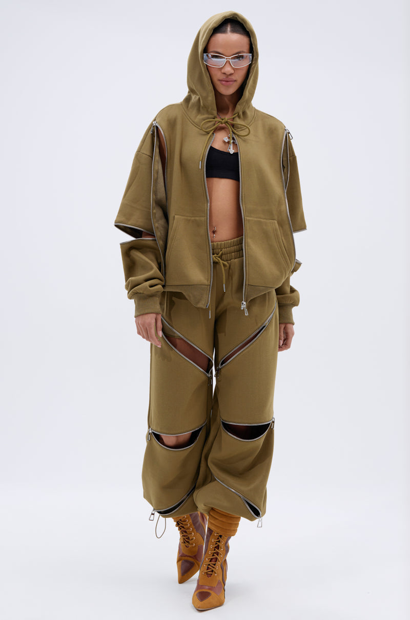 A woman faces forward wearing the CALI MULTI ZIPPER SWEATPANT in olive green, styled with a matching zip hoodie, black bra, brown lace-up boots, sunglasses, and the hood up. Multiple large zippers on the sweatpants reveal her bra.