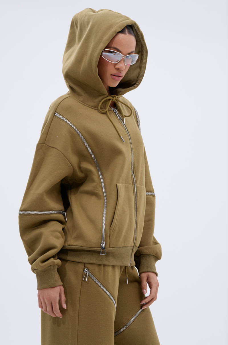 Wearing the CALI MULTI ZIPPER HOODED SWEATSHIRT with bold diagonal zippers, matching pants, and clear futuristic glasses, a person stands against a plain light background.