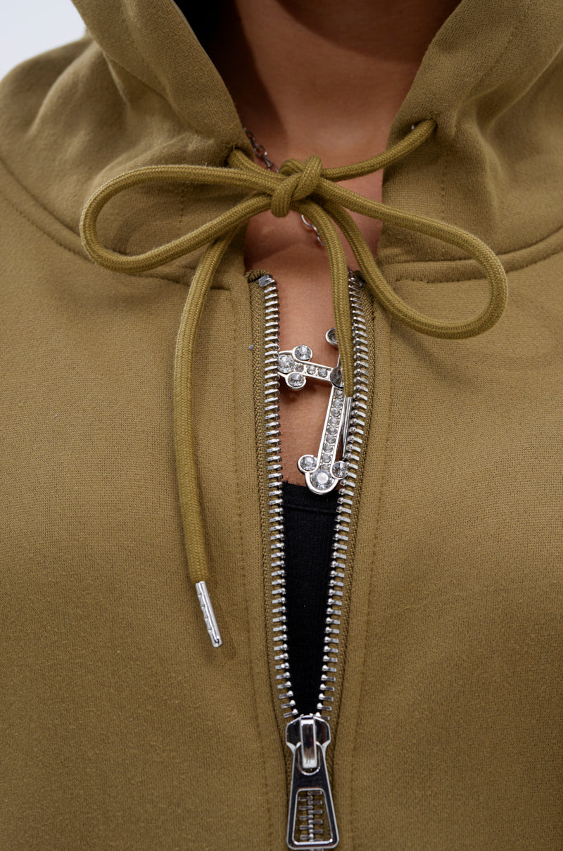 Close-up of a person wearing an olive green CALI MULTI ZIPPER HOODED SWEATSHIRT with a tied drawstring, partially unzipped to show a silver cross necklace and black shirt underneath.