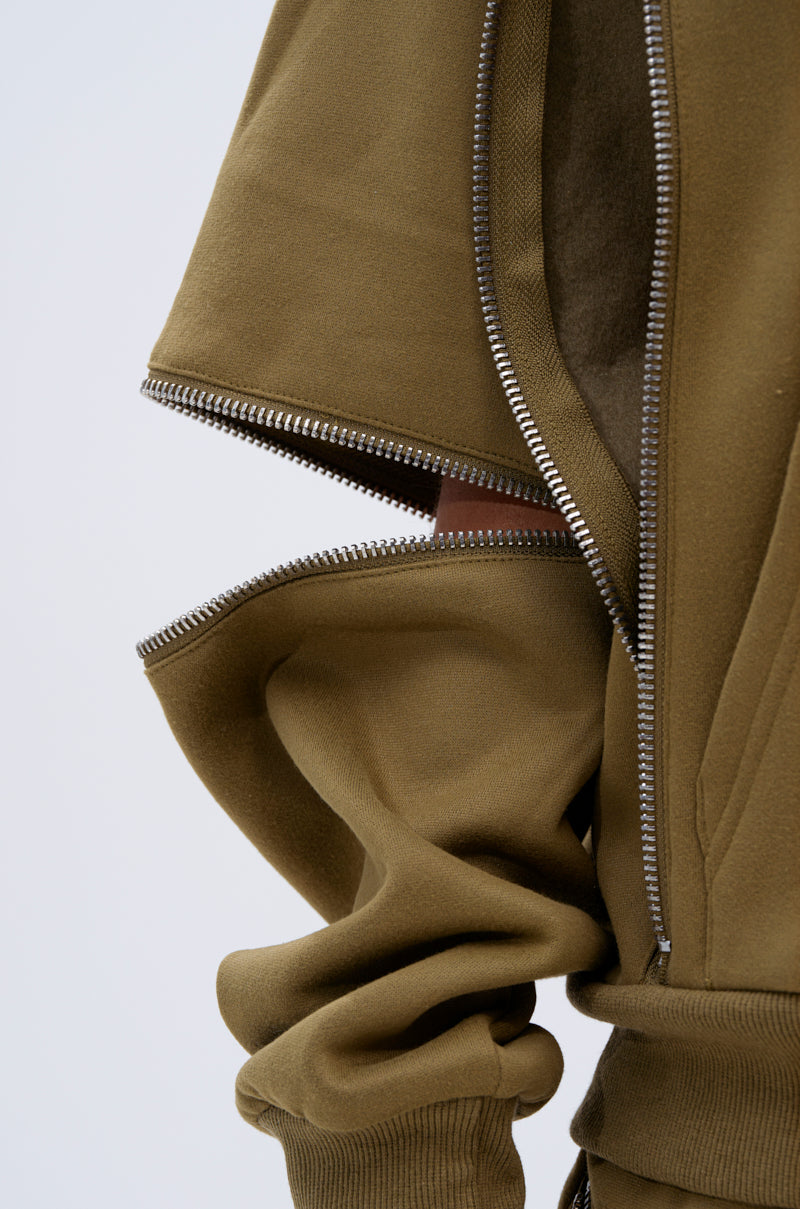 Close-up of an olive green CALI MULTI ZIPPER HOODED SWEATSHIRT featuring multiple silver zippers partially open, revealing layered, soft fleece fabric and forming an abstract, geometric look. Material appears soft with a slightly wrinkled texture.