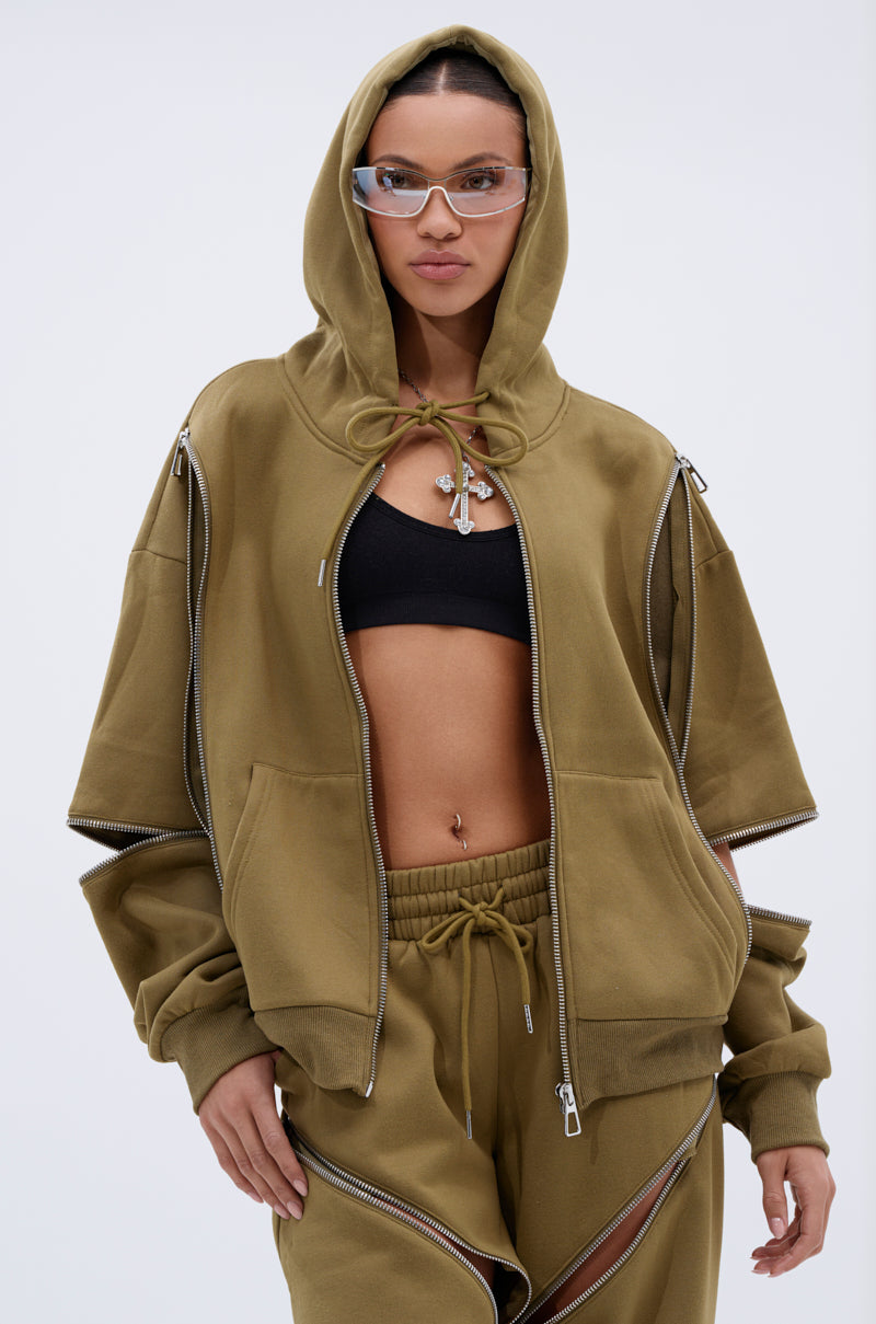 Wearing the CALI MULTI ZIPPER HOODED SWEATSHIRT with wide sleeves, matching pants, a black crop top, clear glasses, and a pendant necklace, a person poses confidently against a plain background.