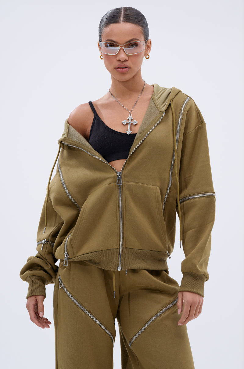 A woman stands confidently in an olive green CALI MULTI ZIPPER HOODED SWEATSHIRT with matching pants over a black sports bra, accessorized with clear glasses, gold hoop earrings, and a silver cross necklace.