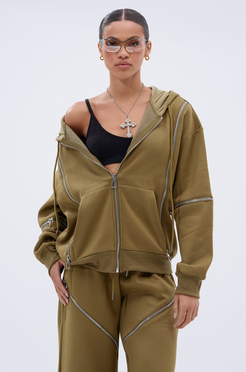 A woman in clear glasses, gold earrings, a cross necklace, a black bra, and an olive green CALI MULTI ZIPPER HOODED SWEATSHIRT with matching pants stands against a plain light background.