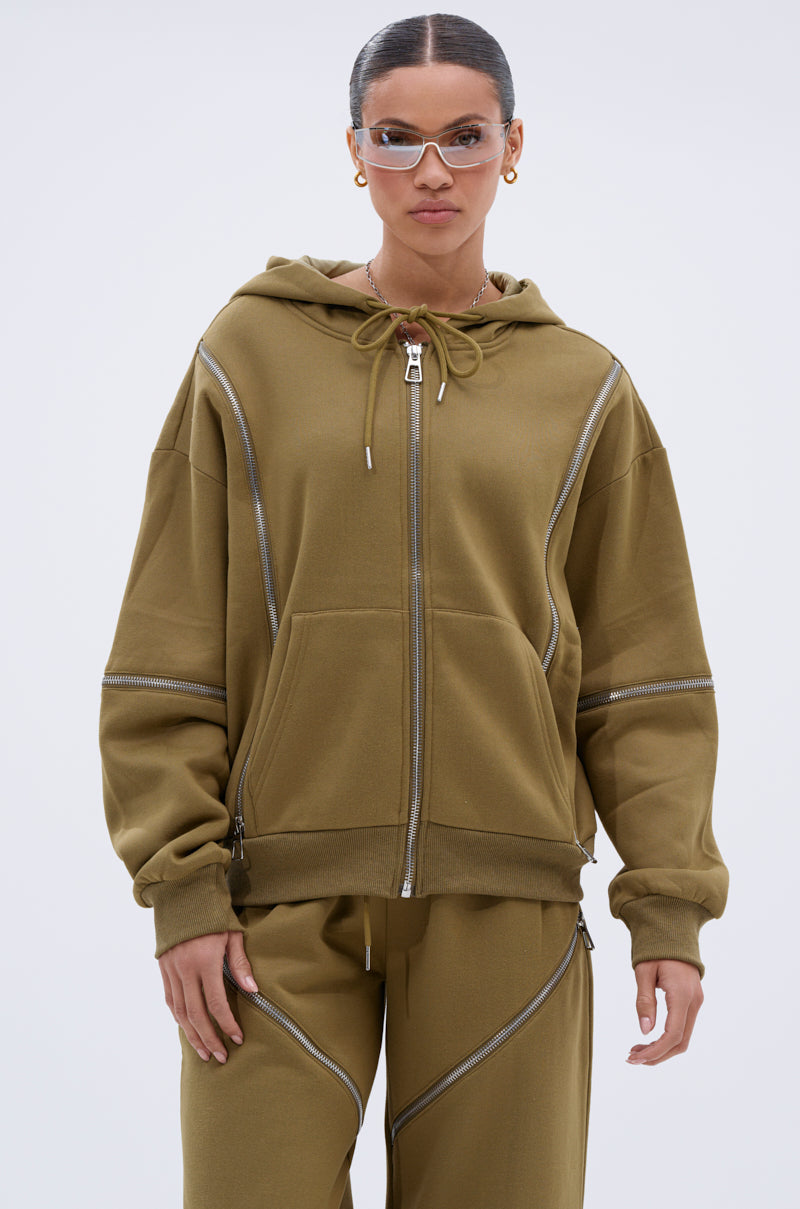 A woman wears the loose-fitting olive green CALI MULTI ZIPPER HOODED SWEATSHIRT with matching zip-detail pants, clear glasses, and gold hoops, standing against a plain light background.