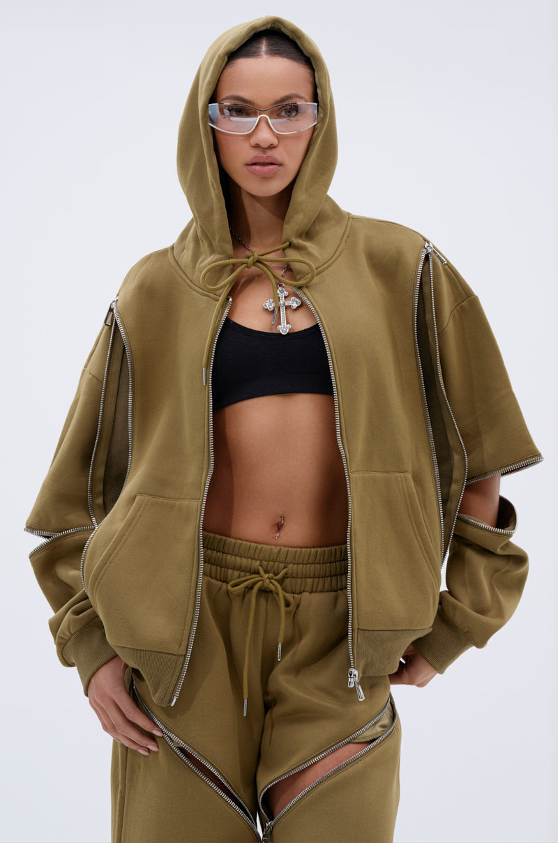 A woman wears the CALI MULTI ZIPPER HOODED SWEATSHIRT in olive green with matching pants from AKIRA Label and a black sports bra. Clear-framed glasses and open zippers on her jacket and pants complete her unique look.