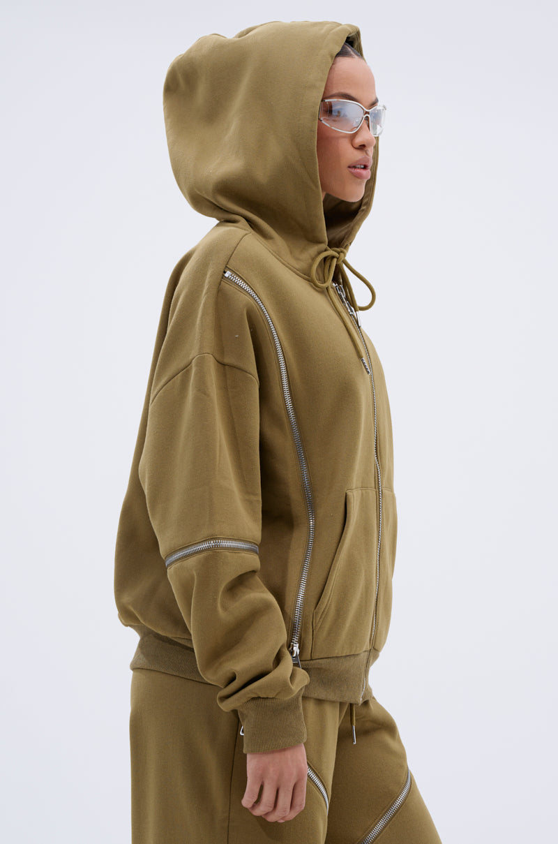 A person wears the CALI MULTI ZIPPER HOODED SWEATSHIRT and matching tan pants with silver zippers and clear protective glasses, standing against a plain light background.