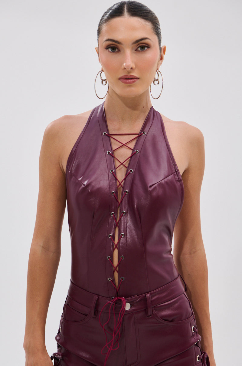 A woman models the SEXY THANG BODYSUIT IN BURGUNDY, a sleeveless faux leather halter with lace-up front, styled with matching pants, hoop earrings, and slicked-back hair against a plain light background. 1