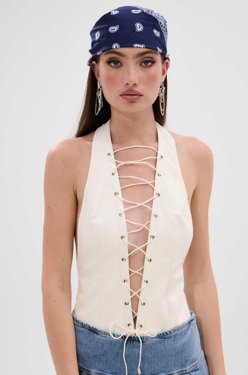 A woman with straight brown hair, wearing hoop earrings and a navy blue bandana, poses in the SEXY THANG BODYSUIT—a sleeveless cream halter style with a lace-up bodice and curve-hugging fit—paired with a denim skirt. She has a neutral expression. 2