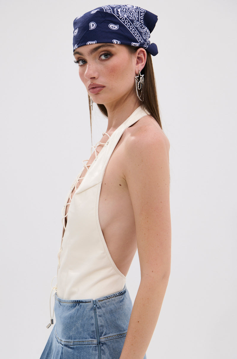 A woman in the SEXY THANG BODYSUIT—a cream halter with a lace-up bodice—paired with a blue denim skirt and navy paisley bandana, glances to the side. She wears statement earrings, has long straight hair, and shows off a curve-hugging silhouette. 3