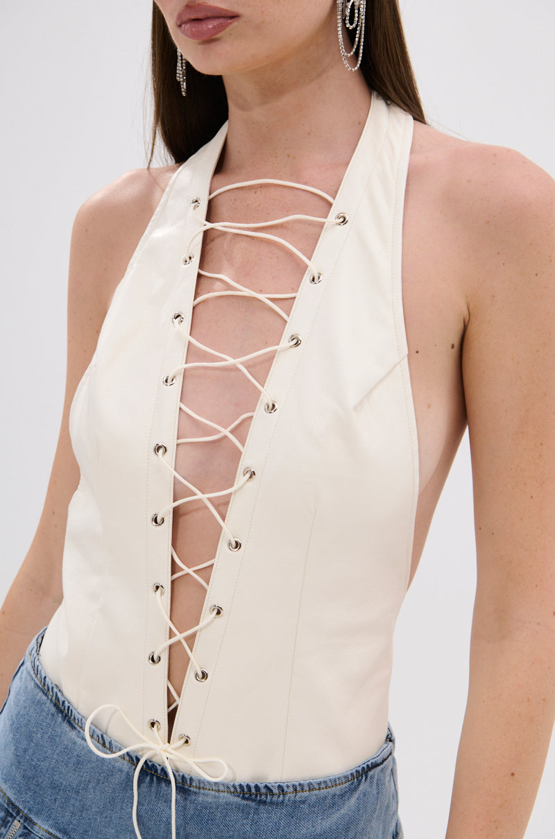 A woman wears the SEXY THANG BODYSUIT, a sleeveless cream halter-neck with lace-up grommet detail and deep V front for a curve-hugging fit, paired with blue denim jeans. She also has long earrings and straight hair. 8
