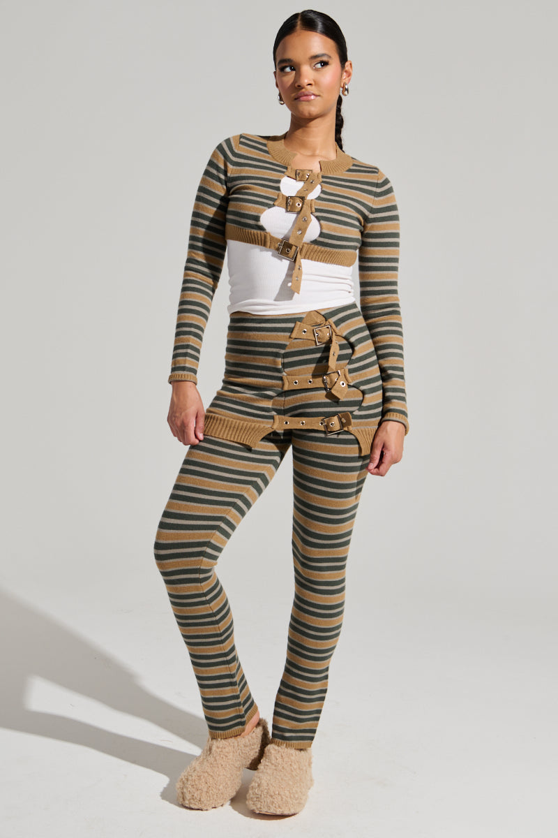 A person stands confidently wearing the SAY LESS FLARE PANT, a green and taupe striped two-piece ensemble that includes a long-sleeved top with distinctive buckle details and matching flared pants. The individual has a neutral expression and fuzzy beige slippers, posed against a plain light gray background.
