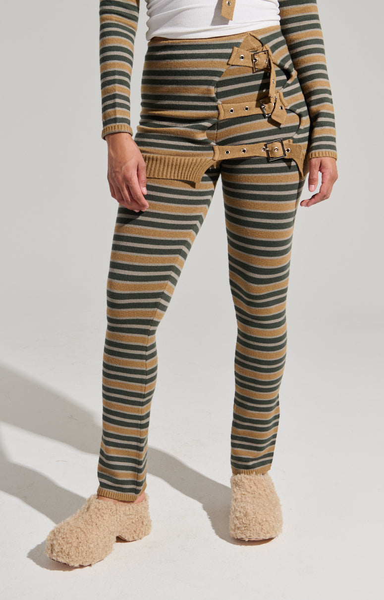 The person is wearing high rise, fitted SAY LESS FLARE PANT with beige and dark green horizontal stripes. The flared pants feature multiple brown leather buckles on one side and a layered waistband. They complete the look with beige, fuzzy slippers.