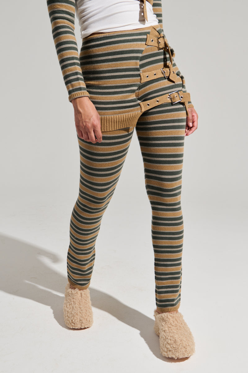 A person wearing SAY LESS FLARE PANT striped in green and tan with multiple brown belts buckled around one thigh, paired with a white long-sleeve shirt and beige fuzzy slippers, stands against a plain background. The stretchy knit pants feature an asymmetrical design and unique fastenings.