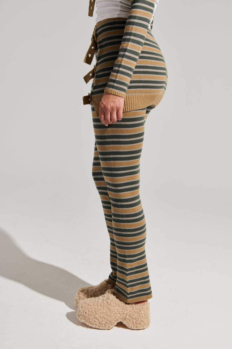 A person stands facing sideways, dressed in SAY LESS FLARE PANT with horizontal stripes in shades of green and beige. They also wear a long-sleeved, fitted shirt with matching stripes and fuzzy beige slippers. The background is plain and light-colored.