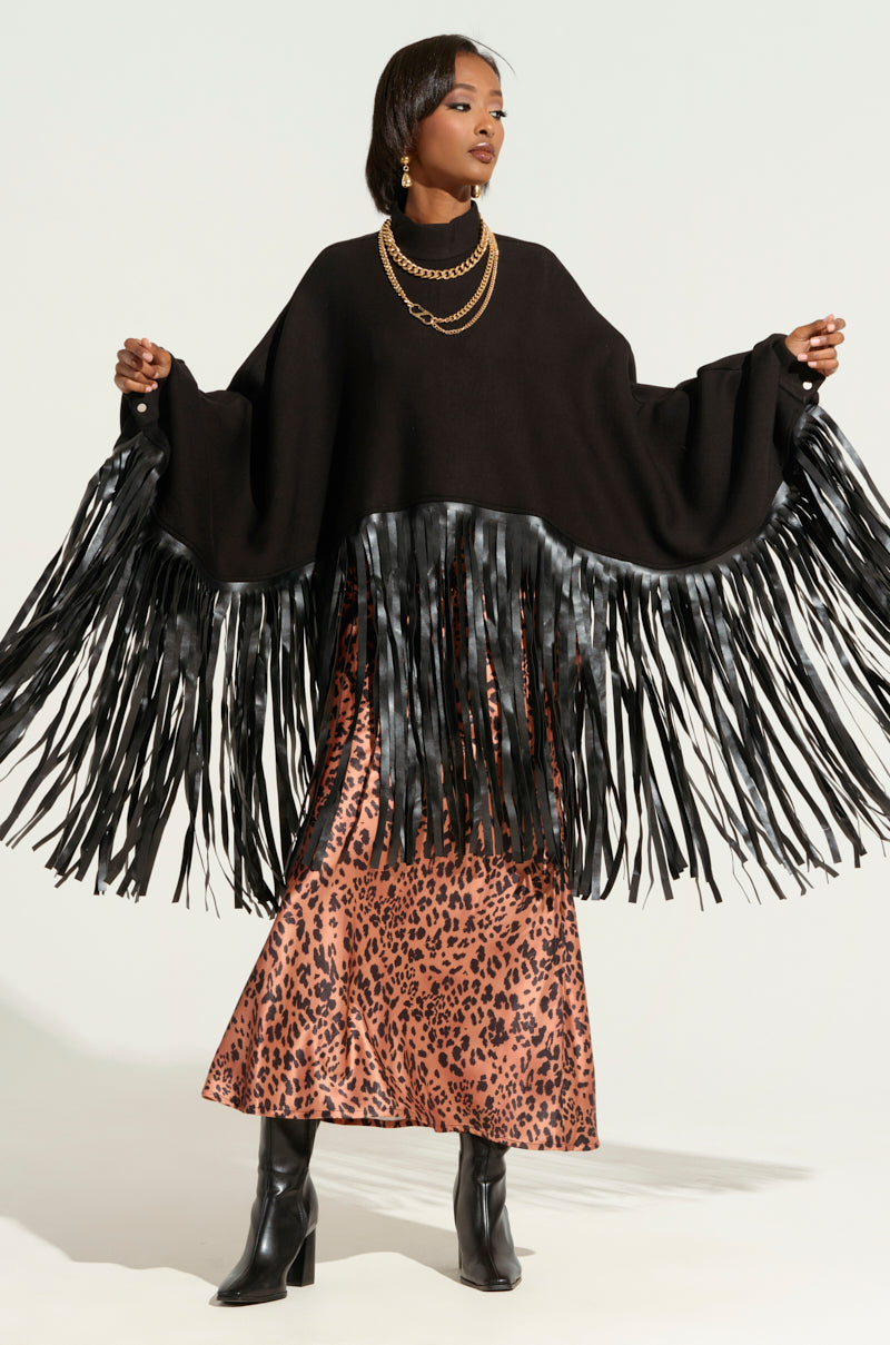 A person poses in the CITY GIRL FAUX LEATHER FRINGE SWEATSHIRT IN BLACK, featuring long faux leather fringe detailing along the sleeves and hem. They wear a leopard-print satin skirt, black ankle boots, and layered gold necklaces from the AKIRA Label. The background is light and neutral.
