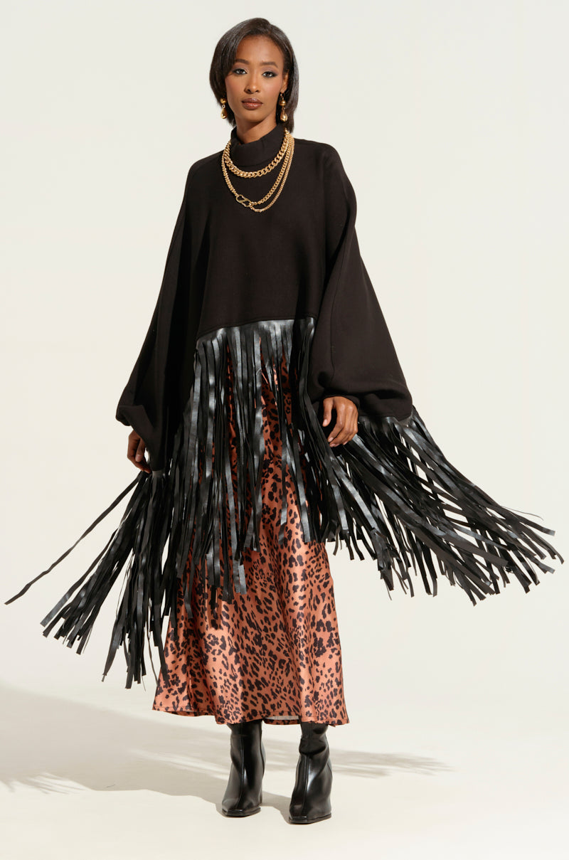 A woman stands confidently, wearing the CITY GIRL FAUX LEATHER FRINGE SWEATSHIRT IN BLACK over a long, leopard print skirt. She accessorizes with layered gold chains and black boots. The background is a plain, light-colored backdrop, highlighting her stylish, eclectic outfit from the AKIRA Label.