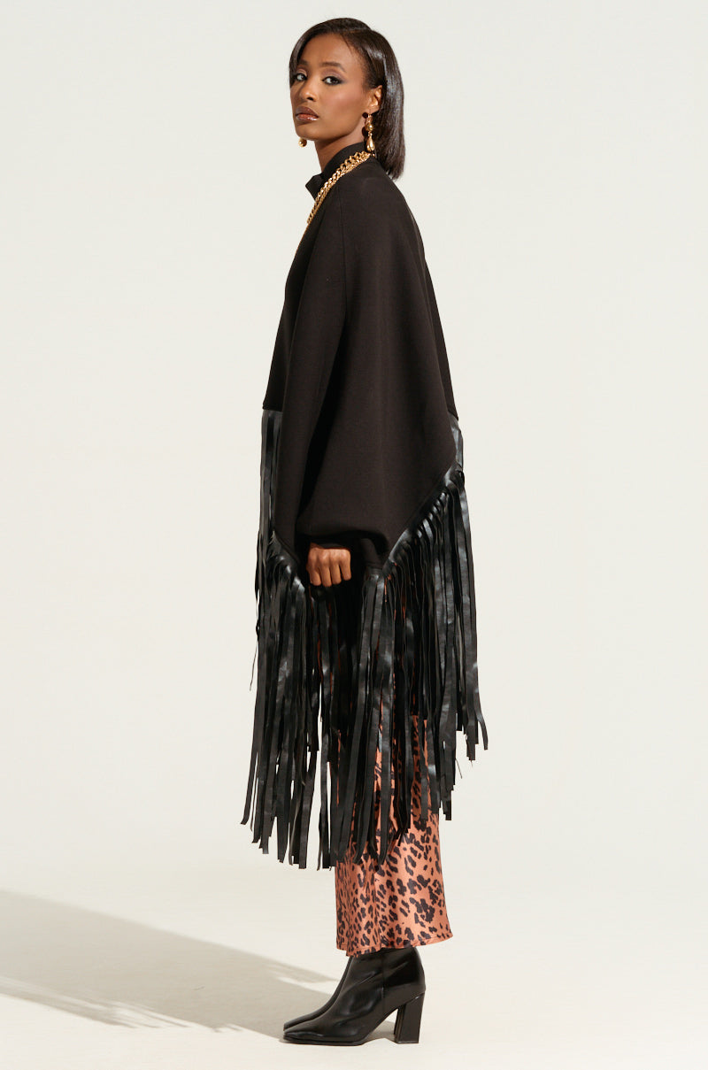 A woman stands in profile wearing the CITY GIRL FAUX LEATHER FRINGE SWEATSHIRT IN BLACK over leopard print pants and black heeled boots. She has straight, shoulder-length hair and accessorizes with a chain necklace and earrings. The background is plain white.