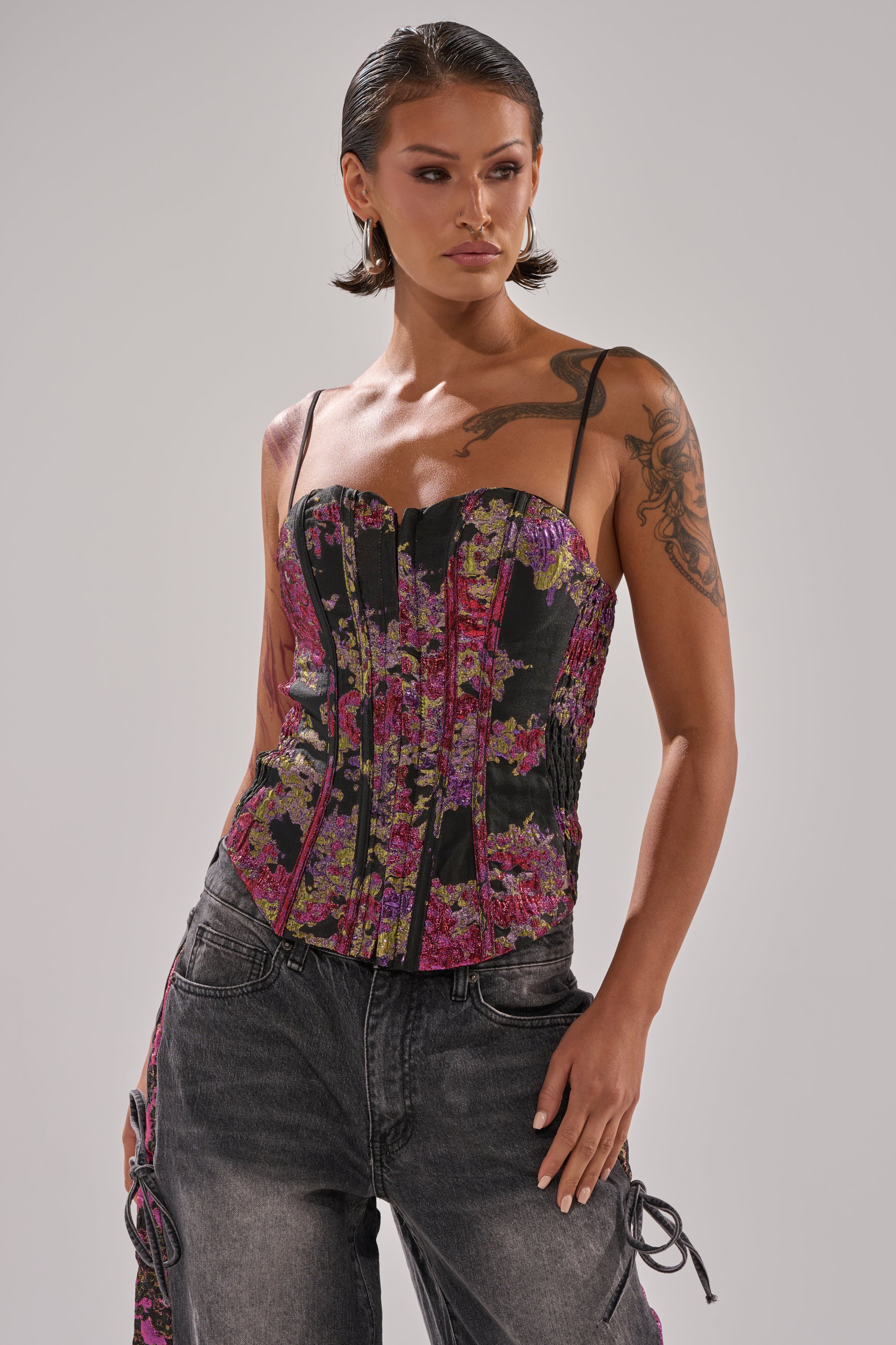 A woman with slicked-back hair wears the LETS PARTY BROCADE CORSET, featuring a sweetheart neckline and thin straps, styled with high-waisted faded black jeans. She shows off arm tattoos and gazes confidently to the side.