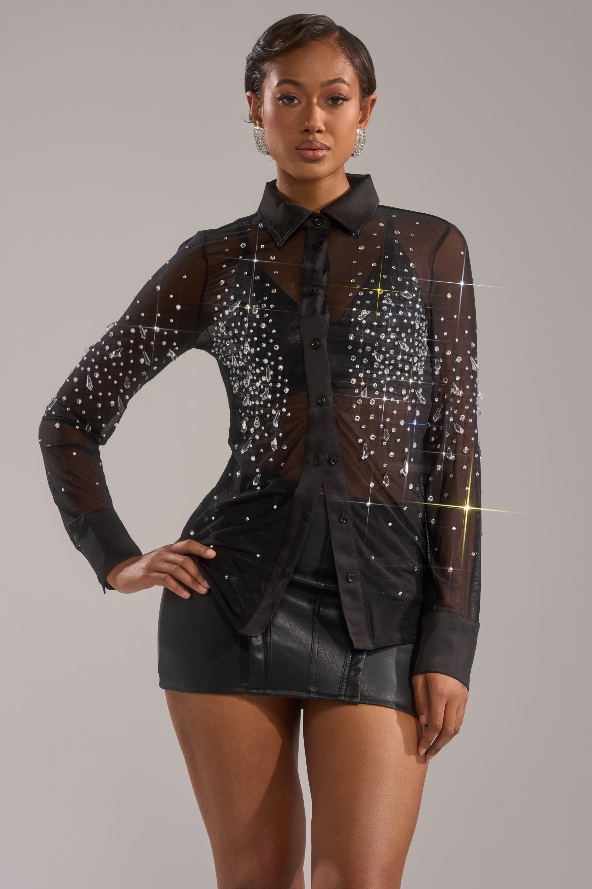 A woman models the BLING BLING MESH BUTTON DOWN with rhinestone details and a black leather mini skirt against a plain light backdrop. 0