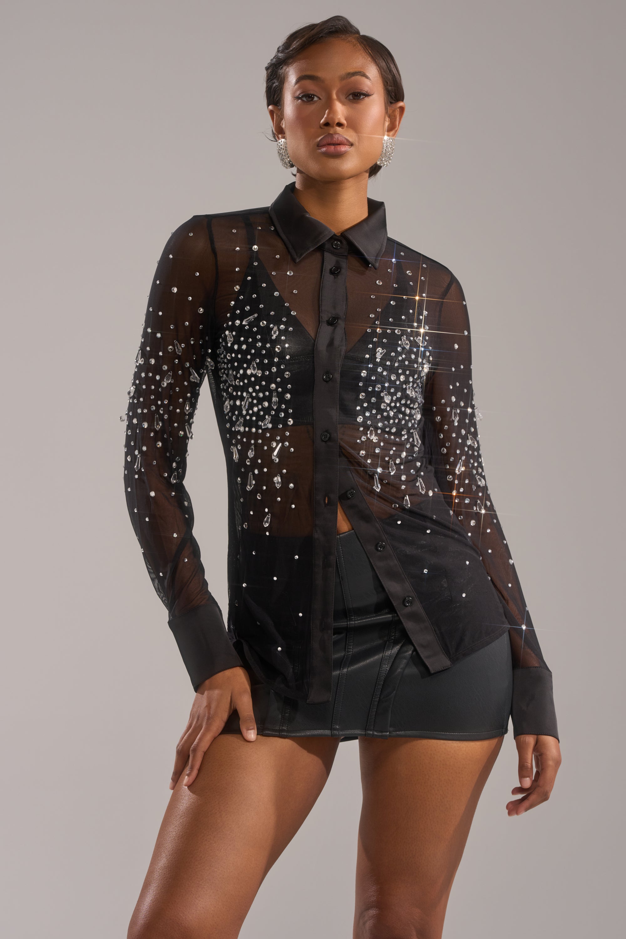 A woman poses confidently in the BLING BLING MESH BUTTON DOWN, a sheer long sleeve shirt adorned with sparkling rhinestones, paired with a black leather mini skirt against a plain gray background. 1