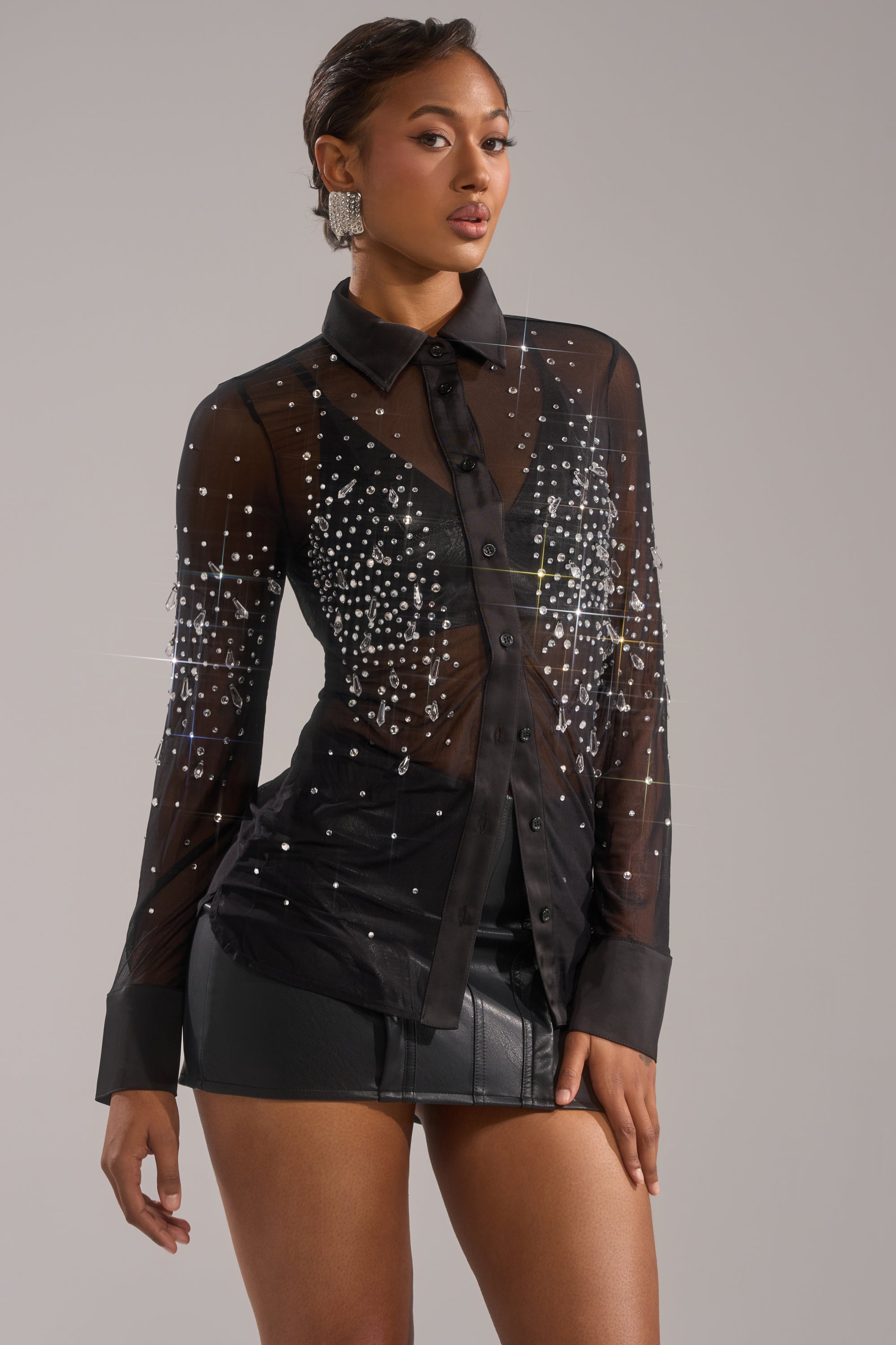 A woman poses in the BLING BLING MESH BUTTON DOWN, a sheer stretchy mesh top adorned with rhinestone embellishments, styled with a short black leather skirt and statement earrings against a plain background. 2