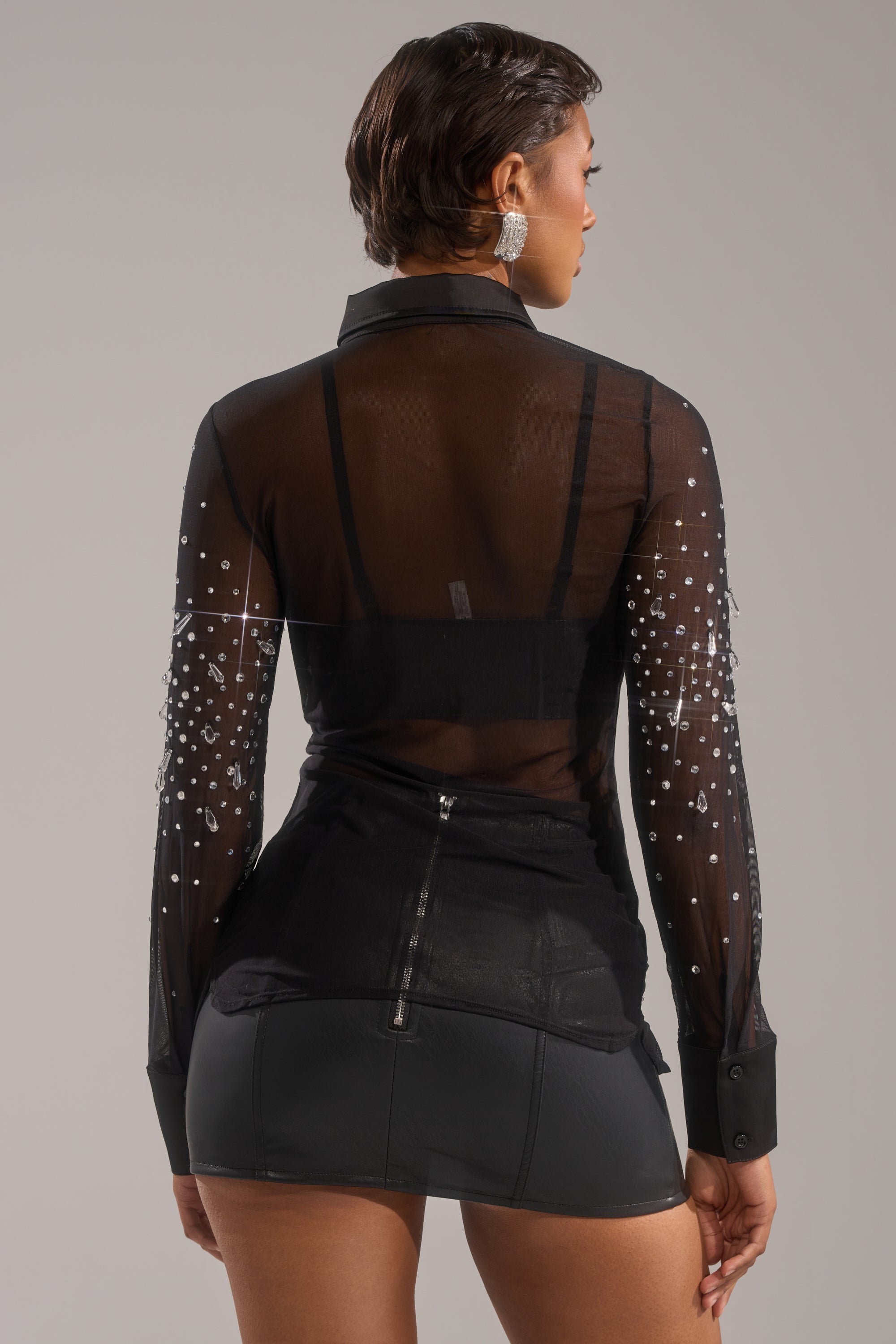 A woman with short hair and large earrings is seen from the back wearing the BLING BLING MESH BUTTON DOWN—a sheer black mesh shirt with rhinestone-embellished sleeves over a black bra, paired with a short black mini skirt. 4