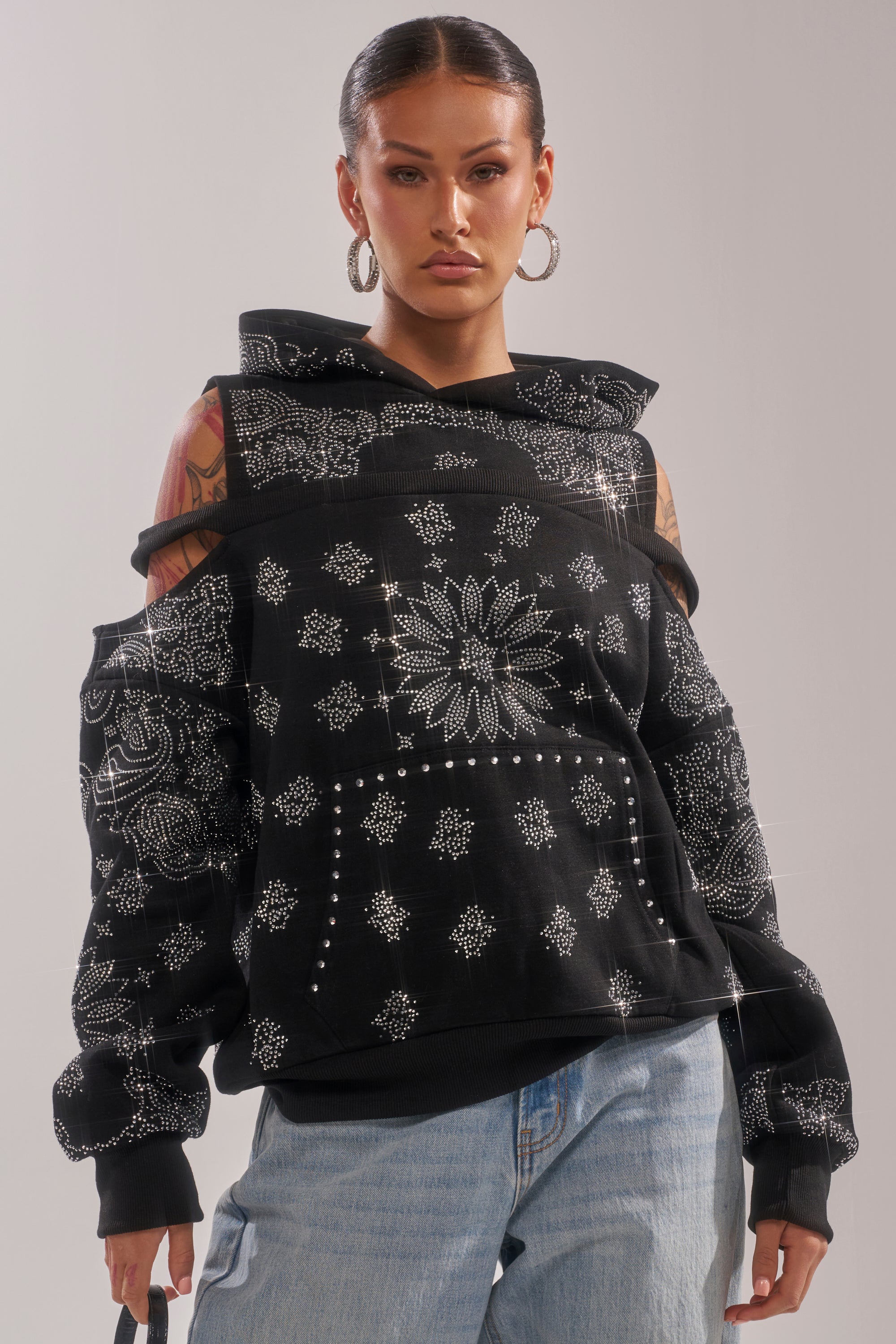A woman in the FEELING FREE SWEATSHIRT—a black hoodie with cut-out shoulders and silver details—pairs it with light blue jeans, hoop earrings, slicked-back hair, and visible arm tattoos, standing against a neutral background. 0