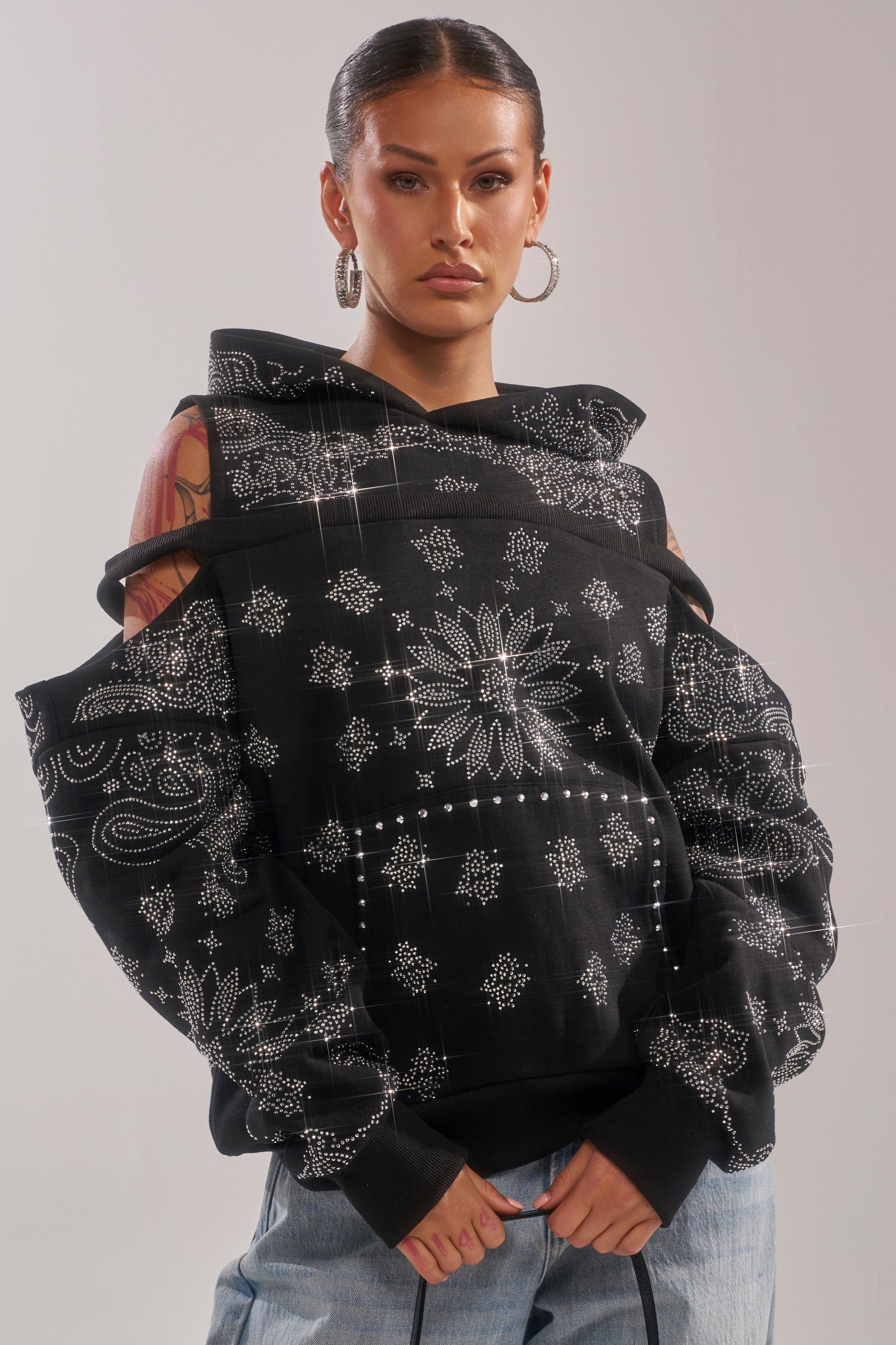A woman with hoop earrings and slicked-back hair wears the FEELING FREE SWEATSHIRT, a black off-shoulder bandana-patterned hoodie, paired with light blue jeans, exuding effortless streetwear style against a plain light background. 1