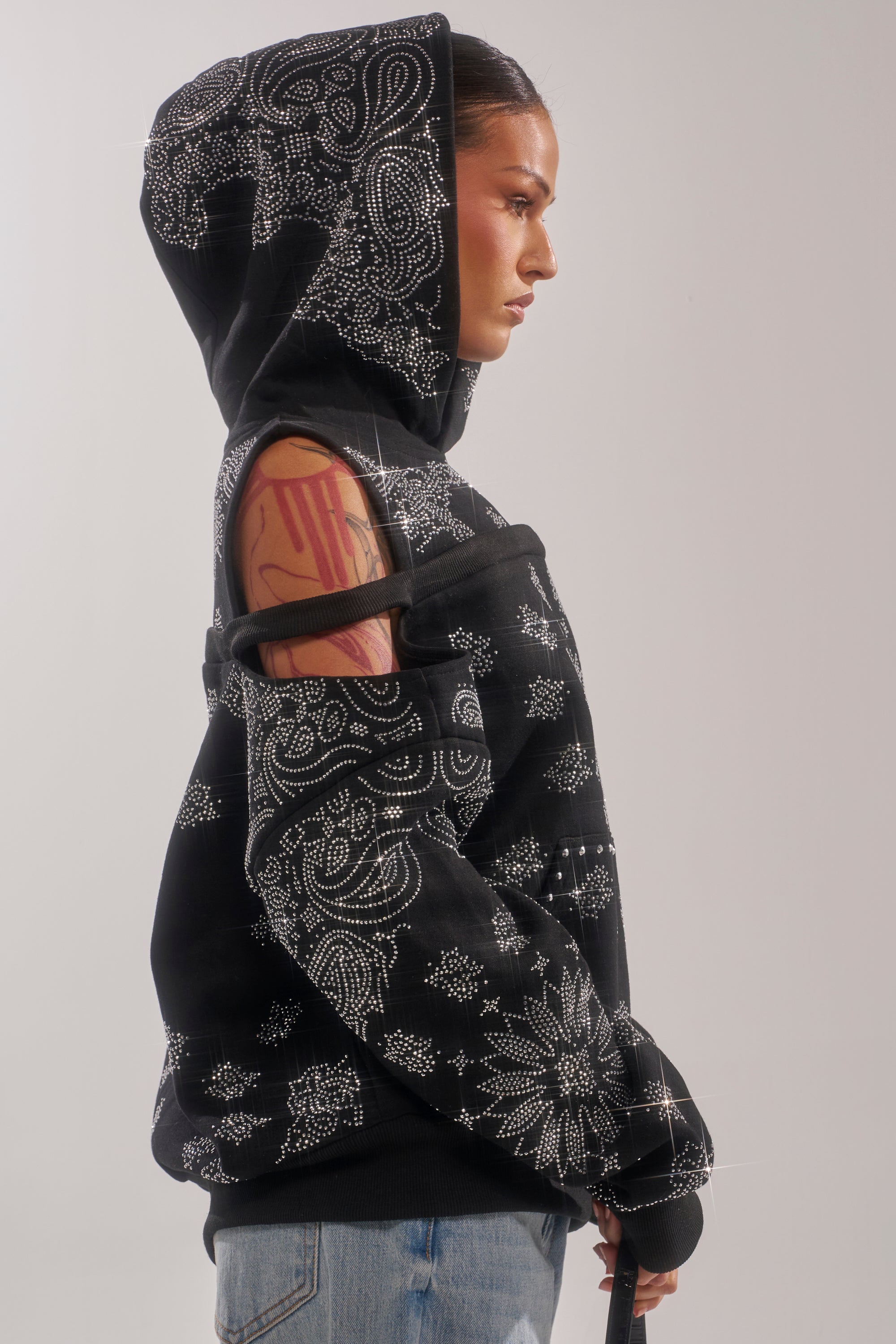 A person in profile wears the FEELING FREE SWEATSHIRT, featuring white ornate patterns and a cut-out shoulder with strap. Their hair is pulled back, pink tattoos are visible on their arm, and they pair it with light blue jeans. 2