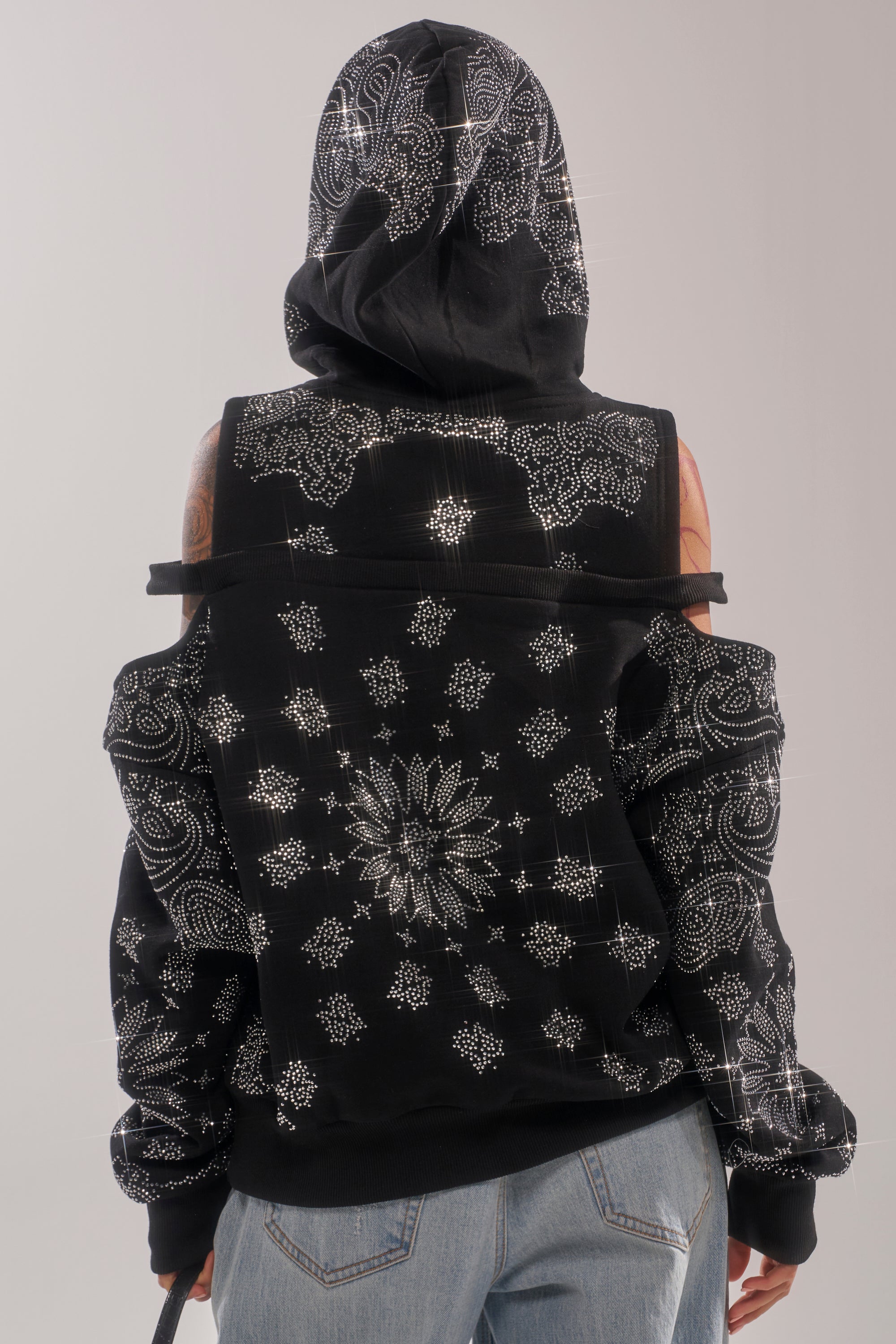 A person shown from the back wears the FEELING FREE SWEATSHIRT—a black, cold-shoulder bandana hoodie with white intricate symmetrical patterns—and light blue jeans. The hood is up, highlighting its unique streetwear style. 3
