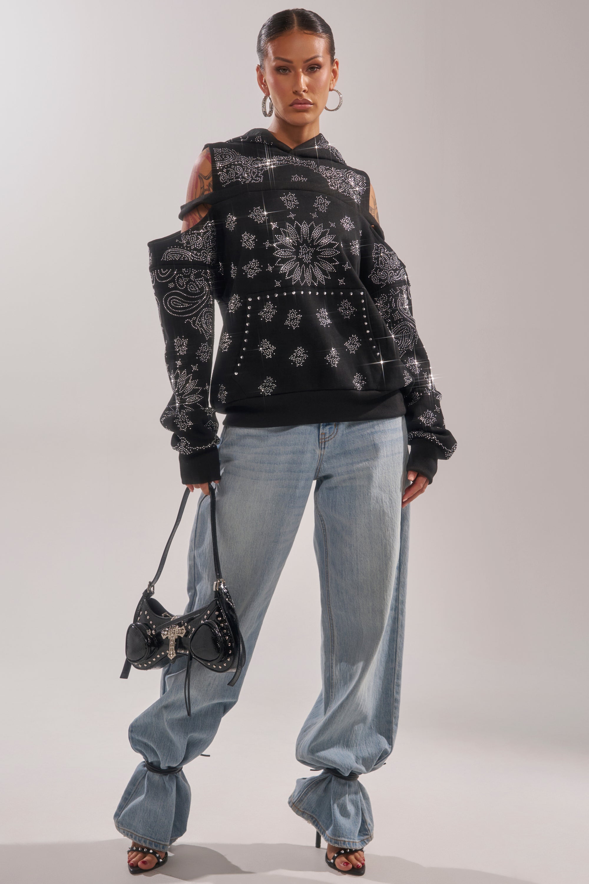 A woman stands confidently in the FEELING FREE SWEATSHIRT—a black bandana-pattern hoodie—paired with loose light blue jeans, heeled sandals, large hoop earrings, and a black studded handbag in a streetwear look against a plain background. 4