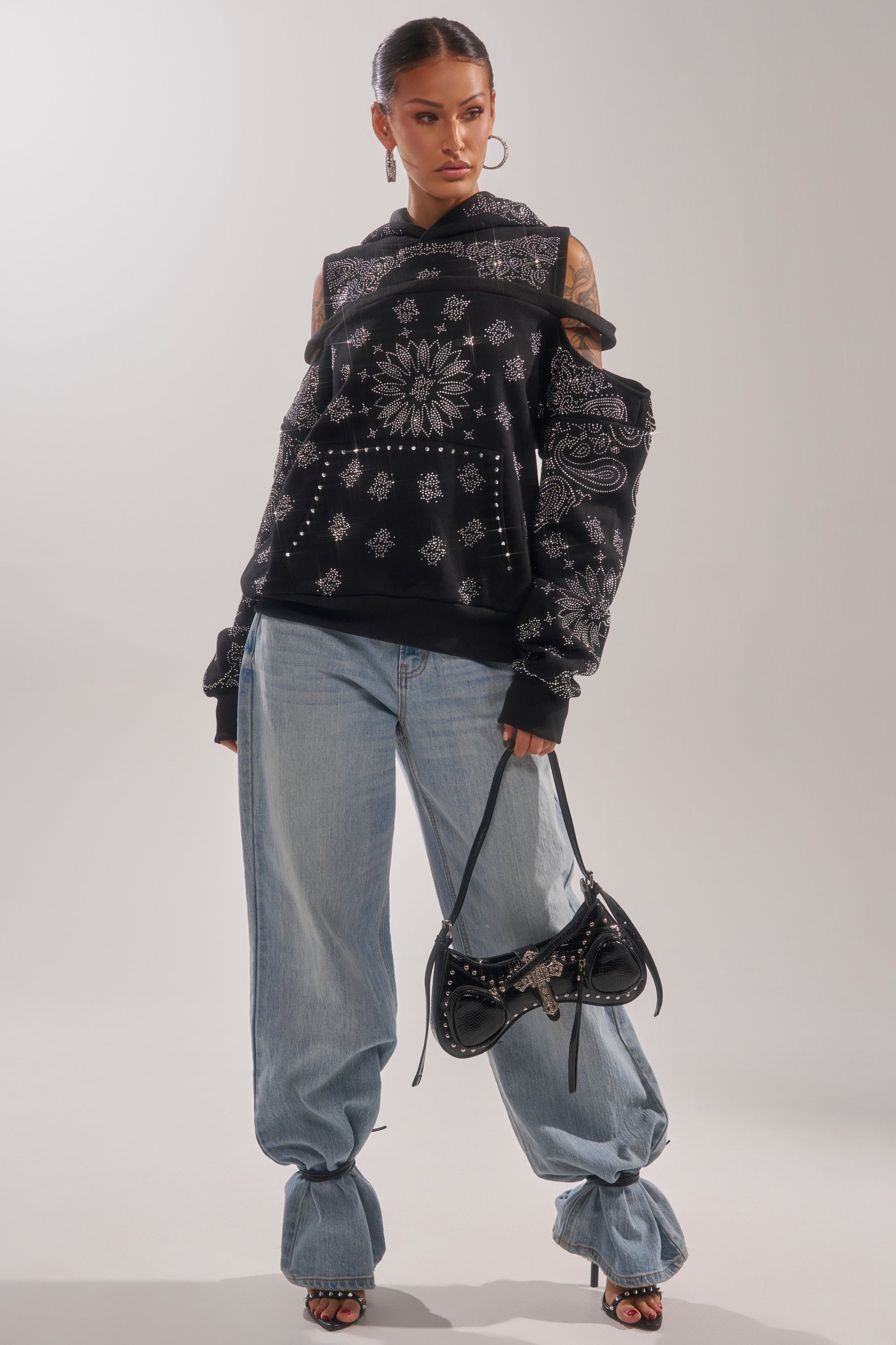 A woman stands confidently in the FEELING FREE SWEATSHIRT with cut-outs, loose light blue jeans, black heeled sandals, and a black handbag. Her hair is slicked back and she completes her streetwear look with hoop earrings. 5