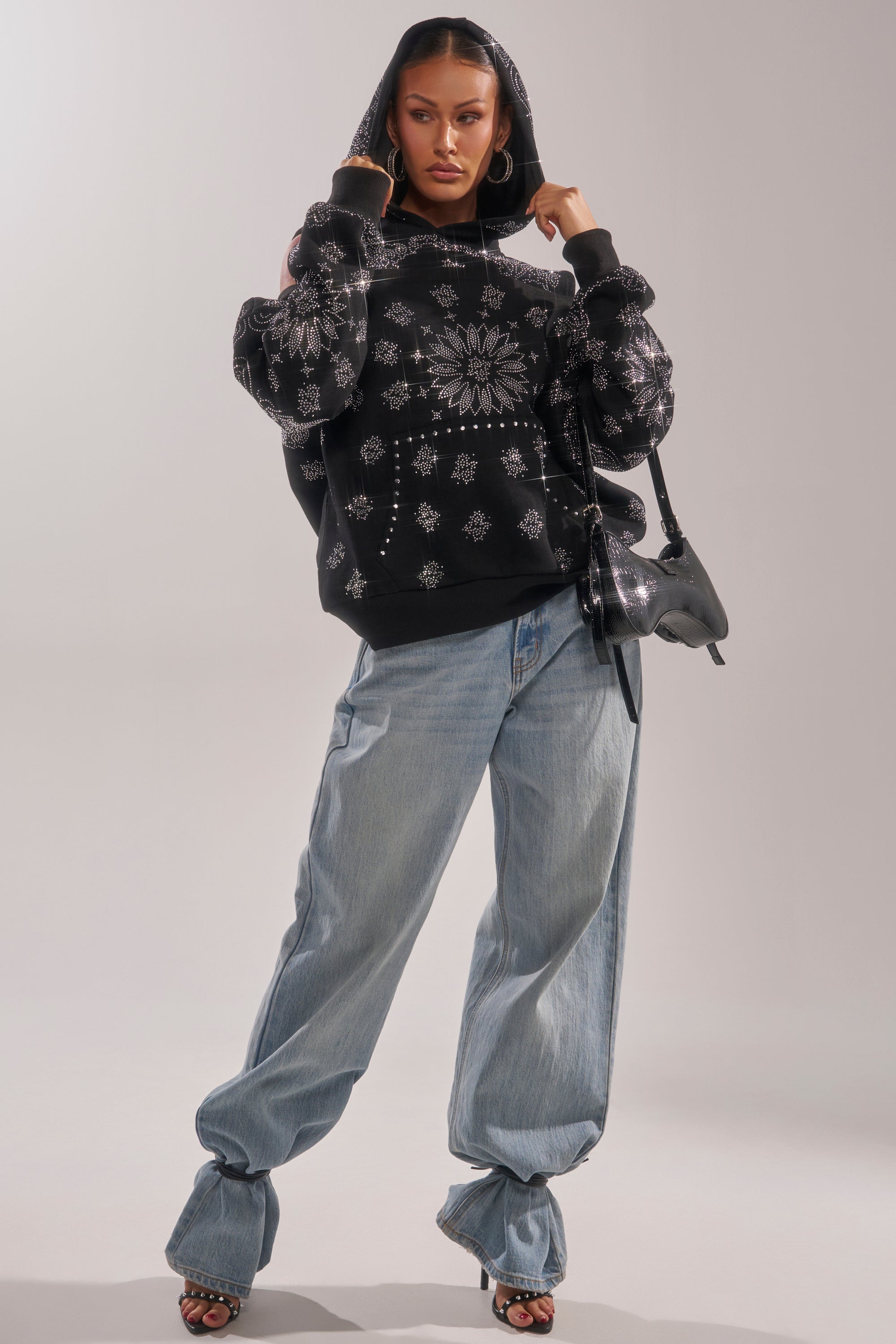 A woman poses confidently in the FEELING FREE SWEATSHIRT, featuring rhinestones and silver snowflakes, paired with light blue baggy jeans, black heeled sandals, hoop earrings, and a shiny black shoulder bag for a bold streetwear look. 6