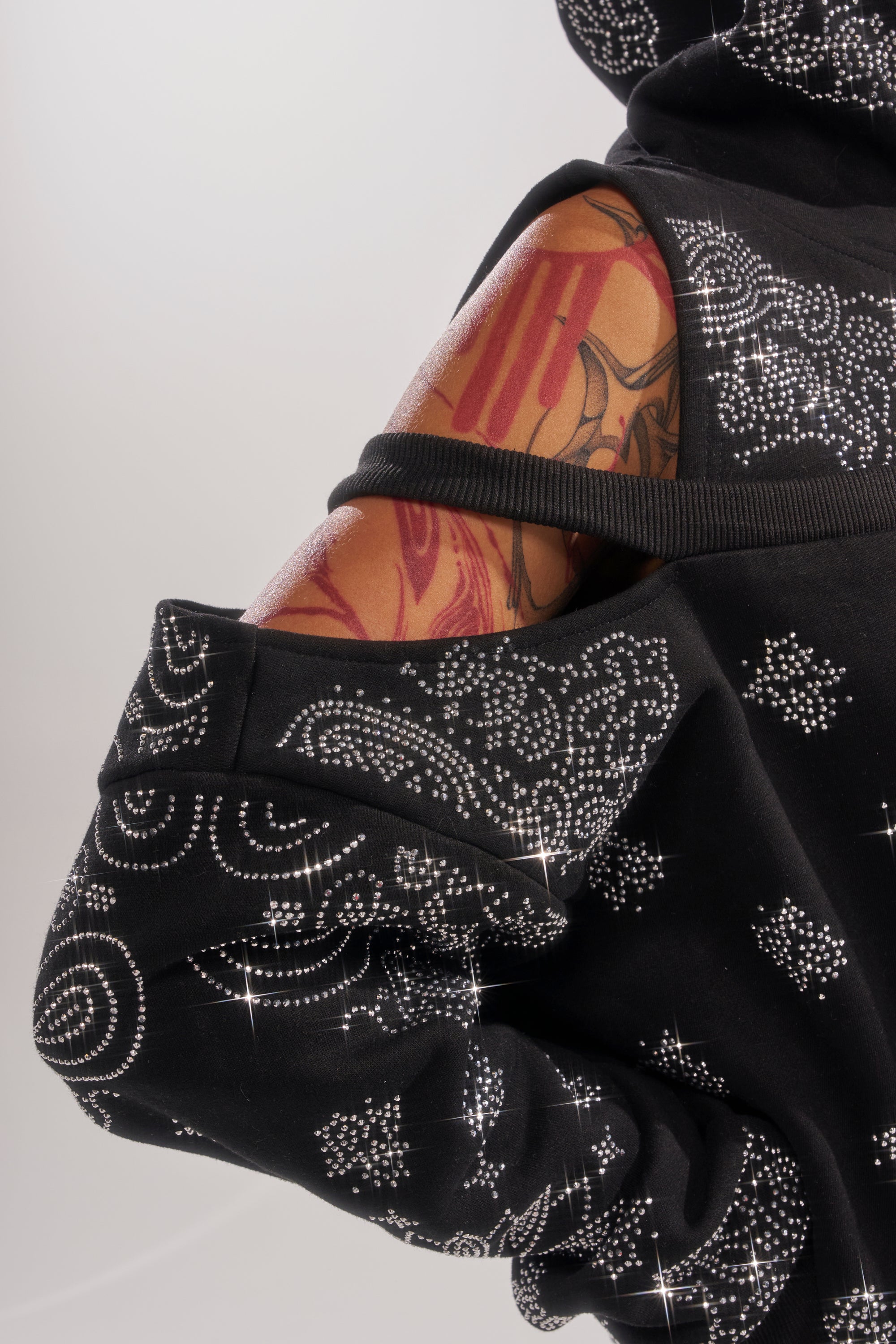 A close-up of a person wearing the FEELING FREE SWEATSHIRT, a black streetwear hoodie with shoulder cutout and sparkling circular rhinestone patterns, showing off a tattooed arm. 7