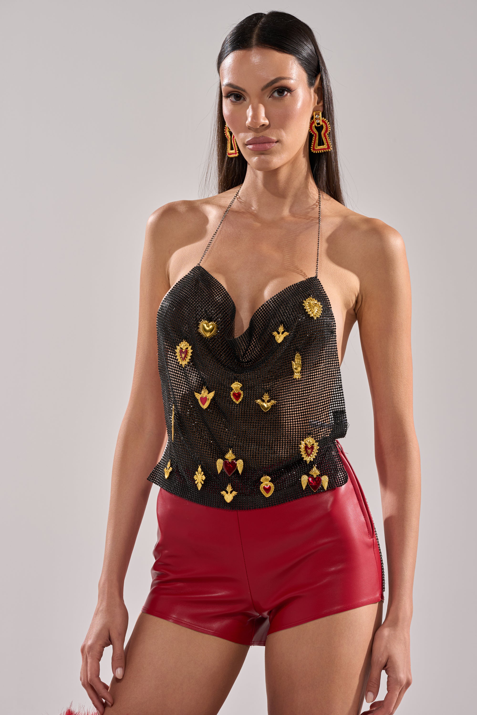 She wears the GILDED MESH HALTER TOP, featuring black mesh with colorful embroidery, paired with shiny red high-waisted shorts and statement red earrings. Her long, straight dark hair complements her confident pose against a plain background. 0
