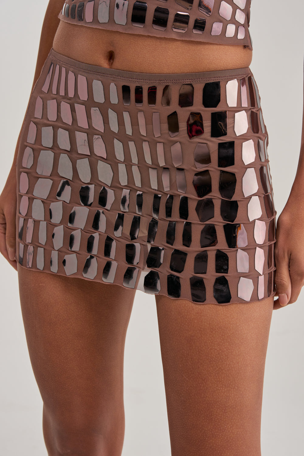 A person wearing the SEE THROUGH ME MINI SKIRT—a fitted brown mesh mini skirt with reflective, square and rectangular embellishments, creating an eye-catching party look.