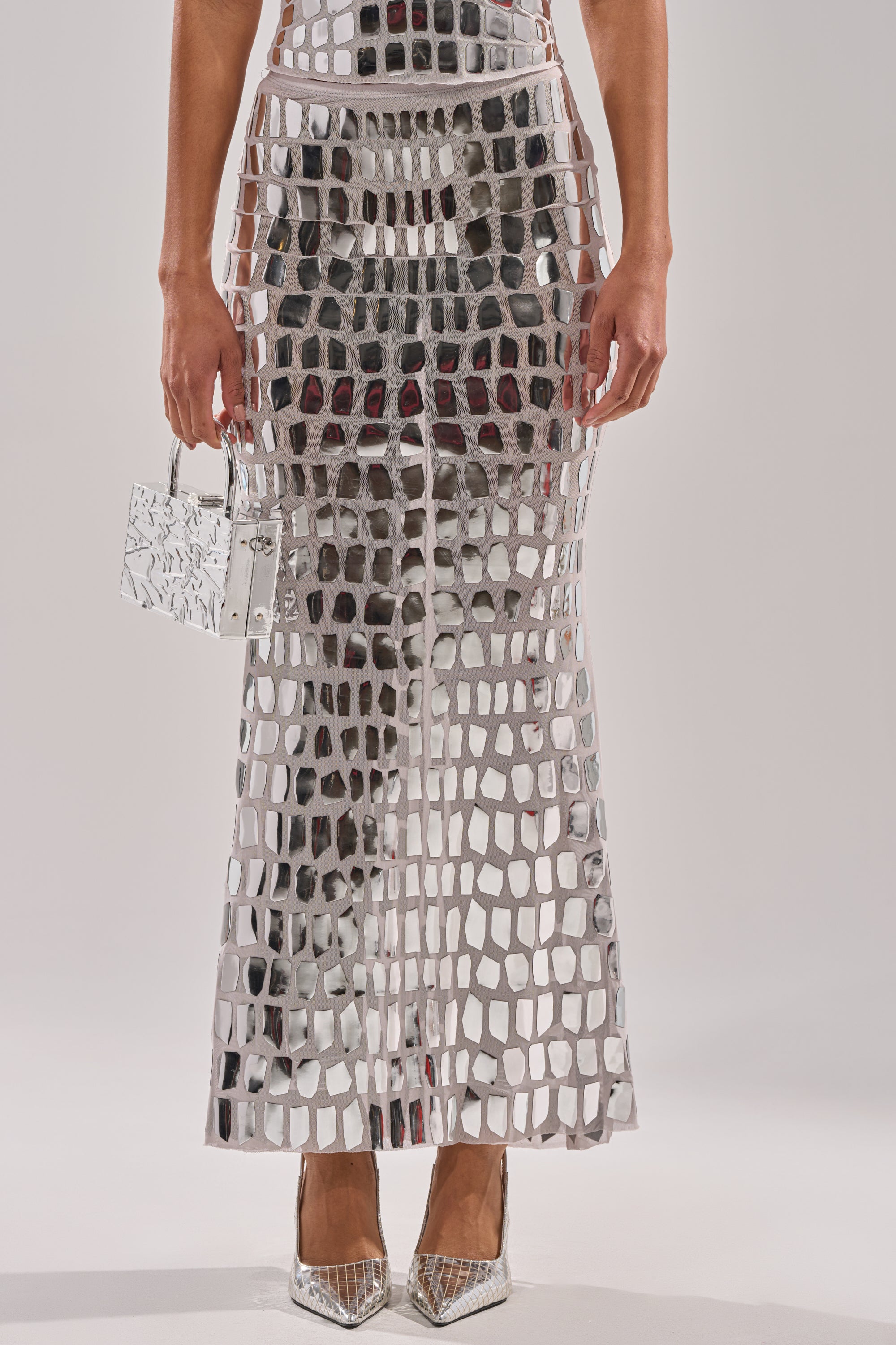 A person wearing the CHAMPAGNE POPPIN MAXI SKIRT—a long, silver, grid-patterned mirrored maxi skirt—holds a small metallic handbag and silver pointed heels against a plain light background.