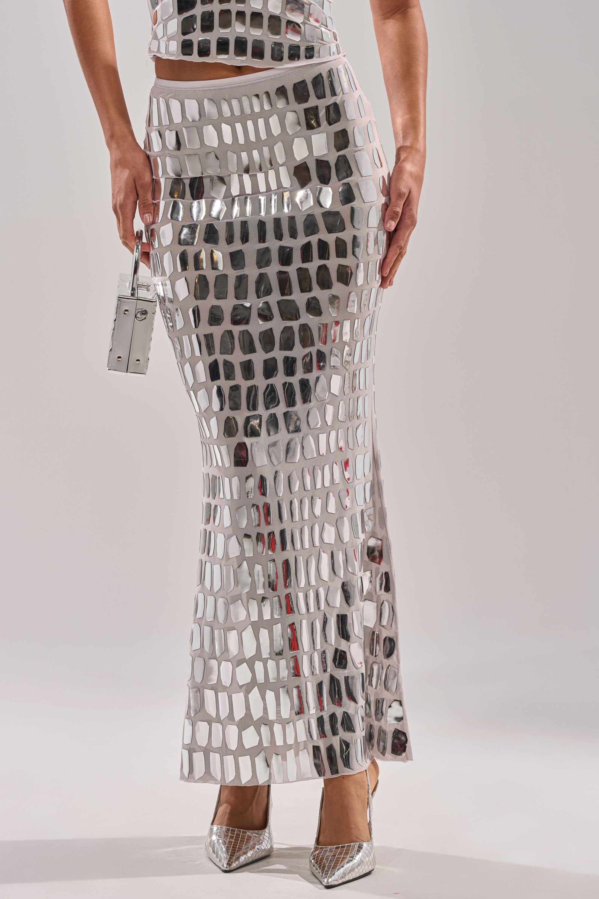Wearing the CHAMPAGNE POPPIN MAXI SKIRT and a matching tile-patterned crop top, accessorized with a silver handbag and pointed heels, creates a bold, reflective look with a striking mirror-like effect.