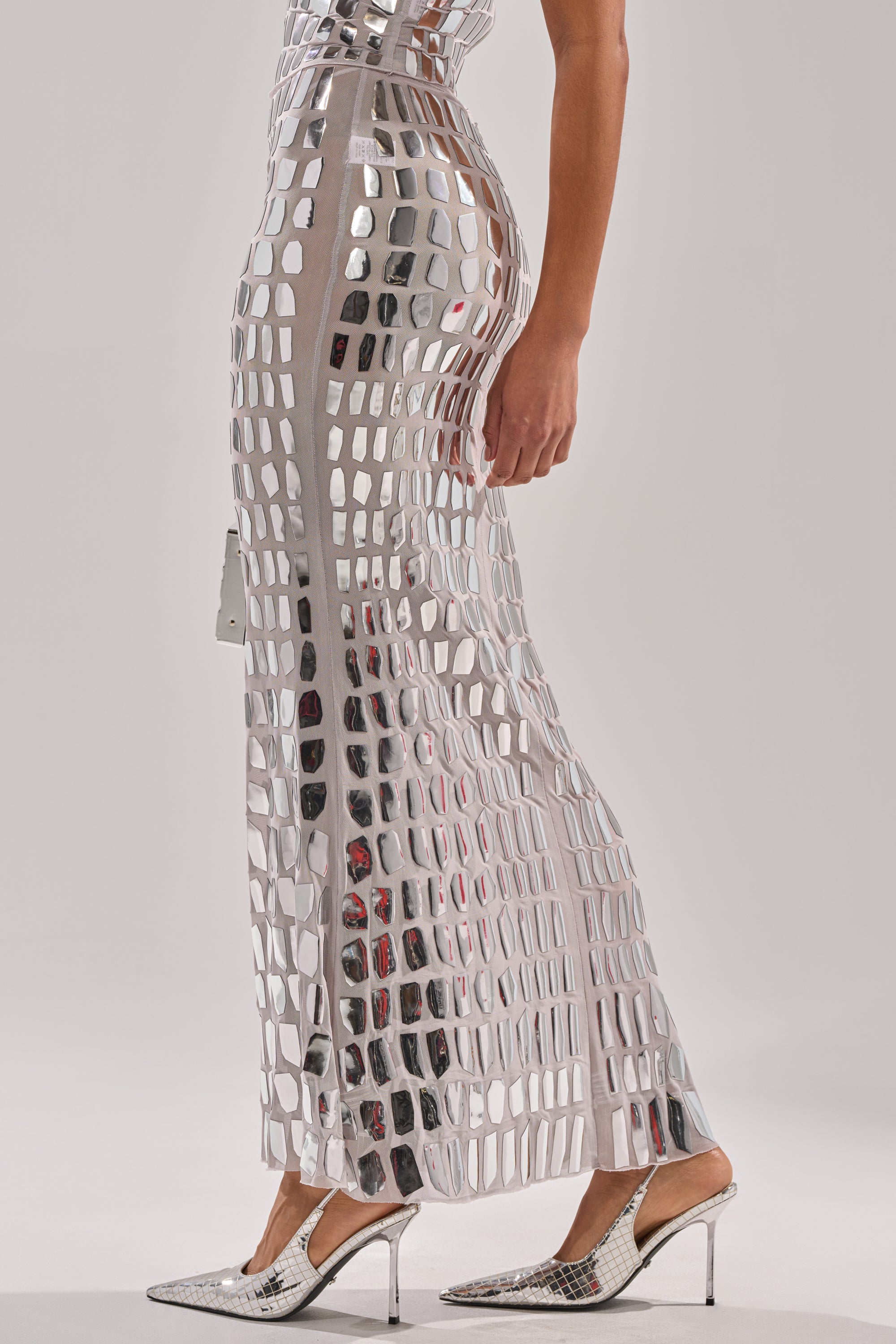 A person wearing the CHAMPAGNE POPPIN MAXI SKIRT, a silver, grid-patterned, reflective statement piece styled with matching high heels, stands against a plain background in a chic, futuristic look.