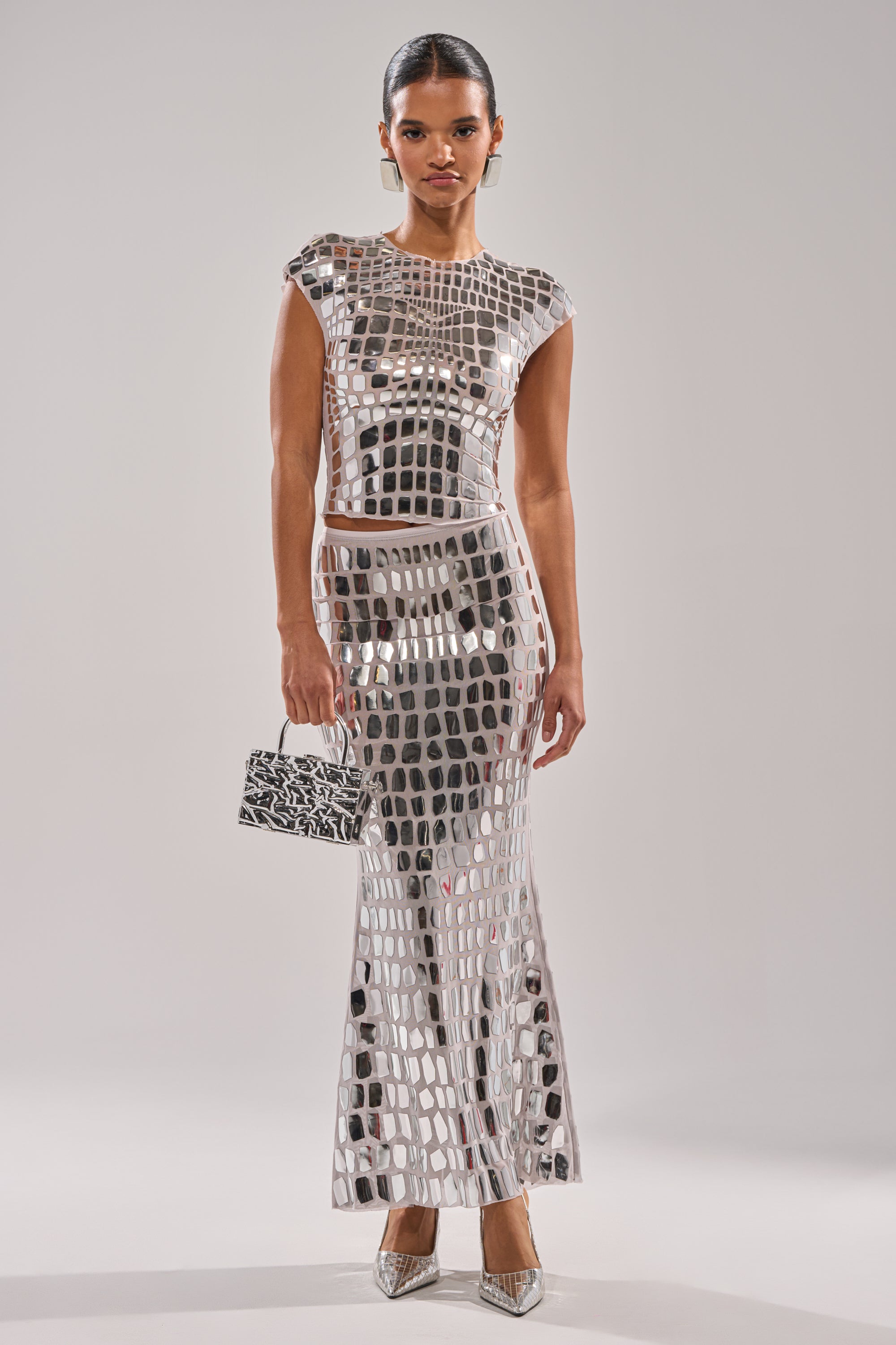 A woman stands against a plain background wearing the CHAMPAGNE POPPIN MAXI SKIRT, a mirrored, floor-length metallic silver skirt with a grid pattern. She holds a small handbag, wears pointed heels, and has her hair slicked back.