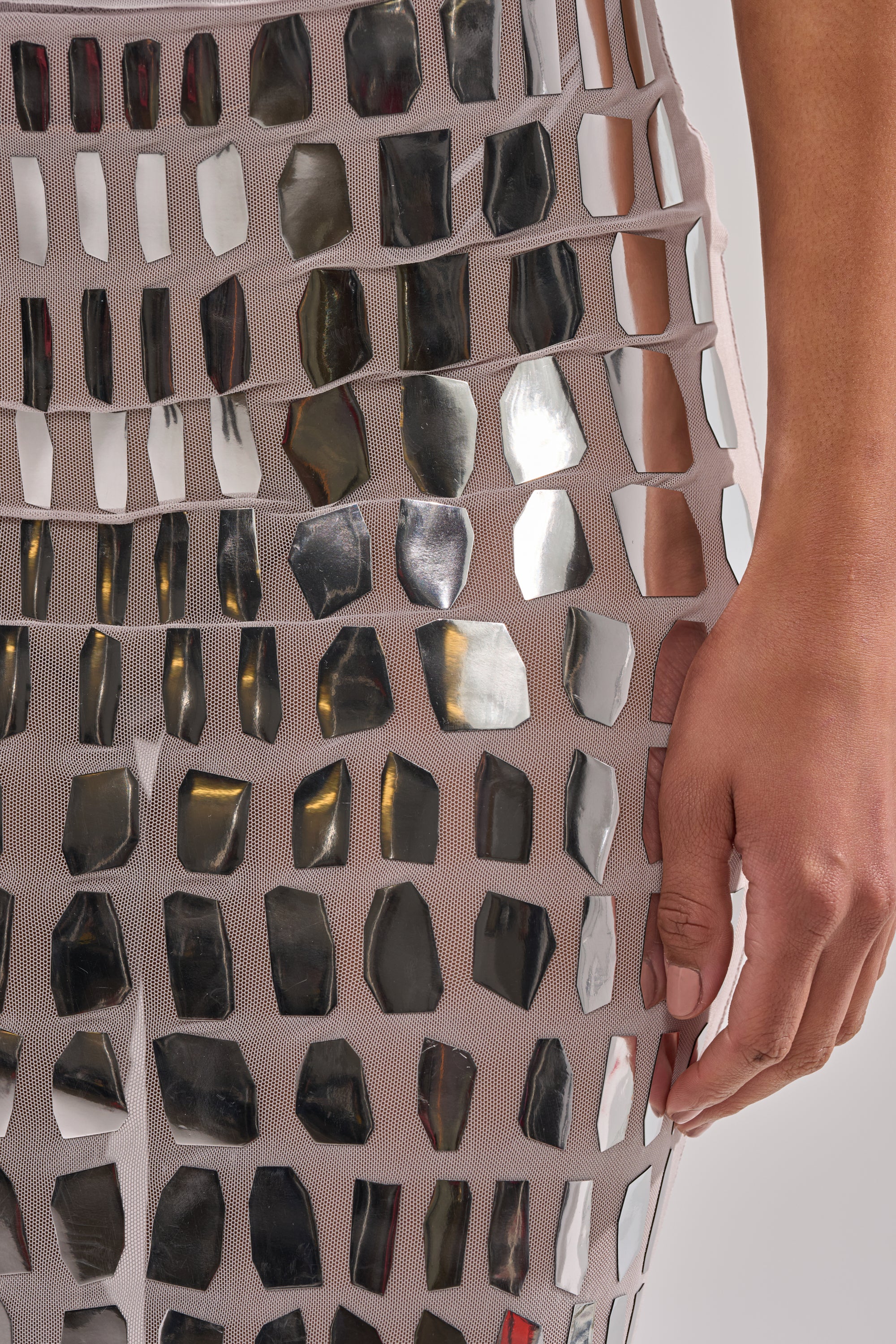 A close-up of a hand on a hip, wearing the CHAMPAGNE POPPIN MAXI SKIRT featuring nude mesh fabric adorned with irregular reflective tiles for a high-shine effect.